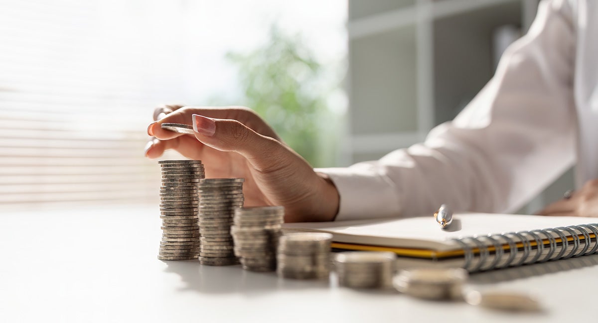 Hand stacking coins into progressively taller piles, with a notebook and pen in the background.