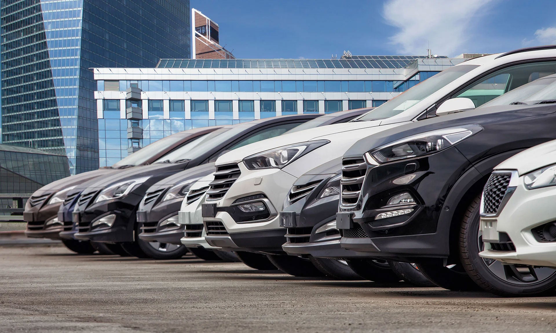 A row of various colored cars, likely SUVs, parked outdoors under a blue sky with clouds and sun glare