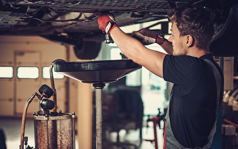 Mechanic in red gloves changing oil under a raised car, with an oil drain pan underneath.