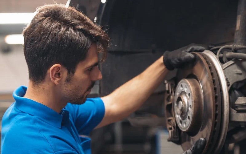 Man in a blue shirt and black gloves adjusting a car's brake rotor in a garage.
